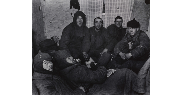 A black-and-white photograph of a group of White men wearing dark, heavy clothing huddled in a small room with plaid curtains over a window.