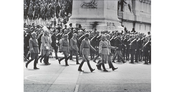 A black-and-white photograph of a military parade, with soldiers lined up to the side watching.