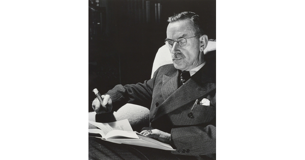 A black-and-white portrait photograph of a seated White man wearing a dark suit and glasses, holding a cigar, and reading a book.