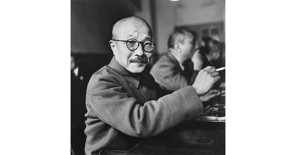 A black-and-white photograph of an Asian man with a shaved head, moustache, and round glasses holding a cigarette.