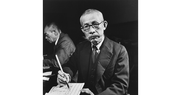 A black-and-white photograph of an Asian man with a shaved head, handlebar moustache, and round glasses writing on paper with a brush and ink.