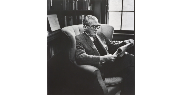 A black-and-white photograph of an older White man in business attire wearing thick, horn-rimmed glasses reading a newspaper in a wingback chair.
