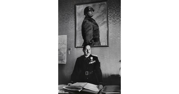 A black-and-white photograph of a White man in military regalia behind a desk stacked with papers above which hangs a portrait of a helmeted man in military dress.