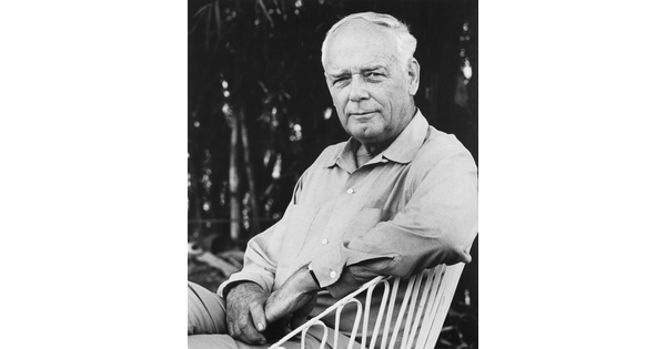 A black-and-white photograph of a seated older, balding White man with a cleft chin wearing a long-sleeved button-down shirt, one arm resting on the back of his chair.