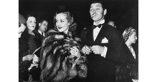 A black-and-white photograph of a White woman wearing a fur coat and a White man with a mustache, smoking a cigarette, wearing a tuxedo.