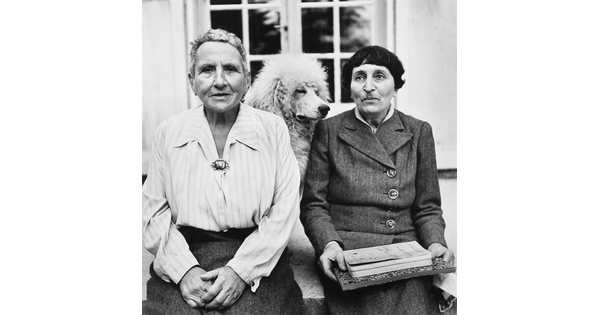 A black-and-white portrait photograph of two older White women seated with a dog in between them.