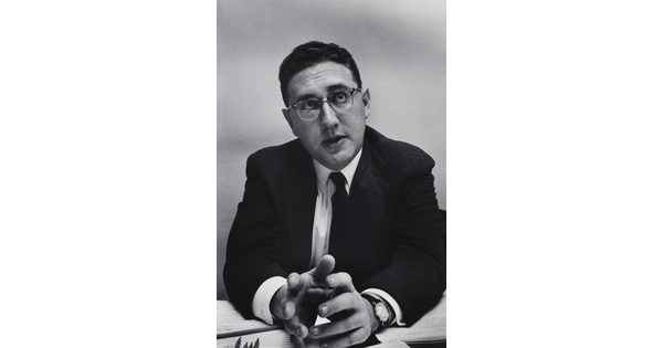 A black-and-white photograph of a White man with short, dark hair in a suit, tie, and glasses leaning forward with his elbows on a desk, gesturing with his hands.
