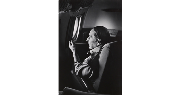 A black-and-white photograph of a White man seated on an airplane smoking a long thin pipe.