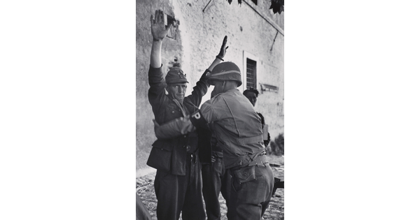 A black-and-white photograph of a White man in military uniform being frisked by a second man in military uniform.