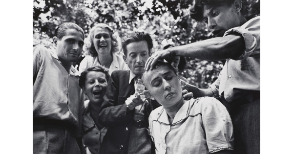 A black-and-white photograph of a seated White woman having her head shaved as laughing and jeering people watch.