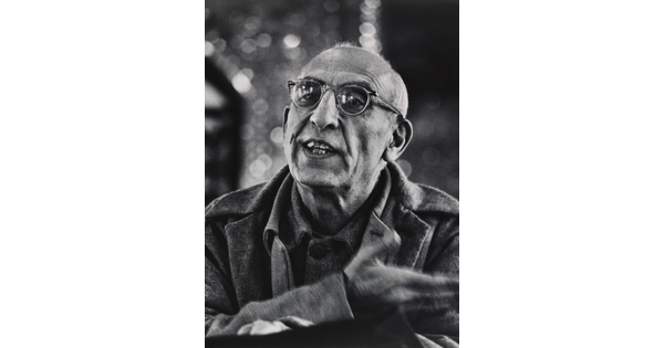 A black-and-white photograph of a balding, medium-skinned man wearing glasses talking and gesturing with his hand.