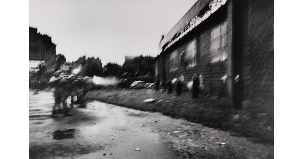 A blurry black-and-white photograph of soldiers on the left shooting rifles at a line of people standing against a wall on the right.