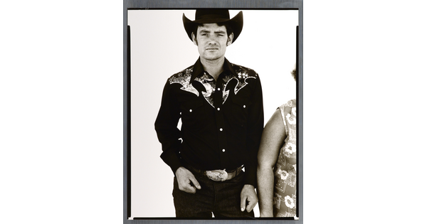 A black-and-white portrait photograph of a White man wearing a dark button-down shirt with Western details, a large belt buckle, and a cowboy hat.