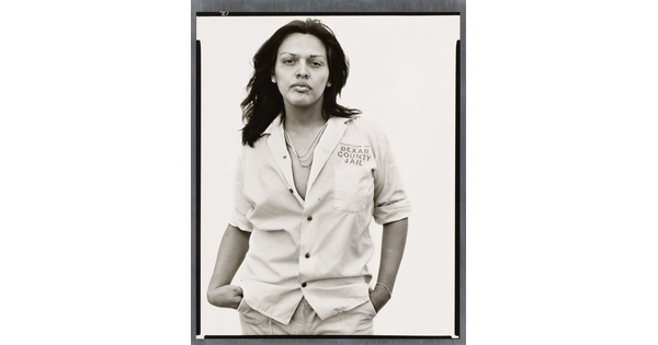 A black-and-white photograph of a medium-skinned person, with long hair wearing a light, partially unbuttoned shirt with text "Property of Bexar County Jail" above the breast pocket.