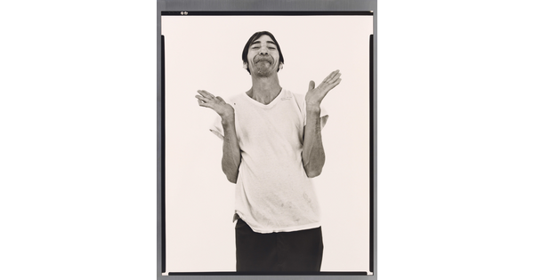 A black-and-white portrait photograph of a medium-skinned man with short hair, light facial hair, wearing a white t-shirt, dark pants, and hands open palmed and raised to shoulders.