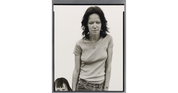 A black-and-white portrait photograph of a weary-looking White woman standing beside a child whose head and body is mostly out of frame.
