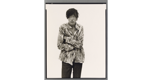 A black-and-white portrait photograph of a light-skinned man with dark hair that hangs into his face standing with arms crossed.
