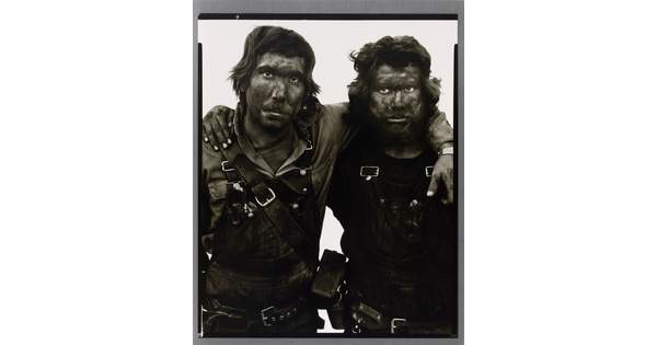 A black-and-white portrait photograph of two men covered in dark coal dust, both wearing dirty overalls, with their arms around one another.