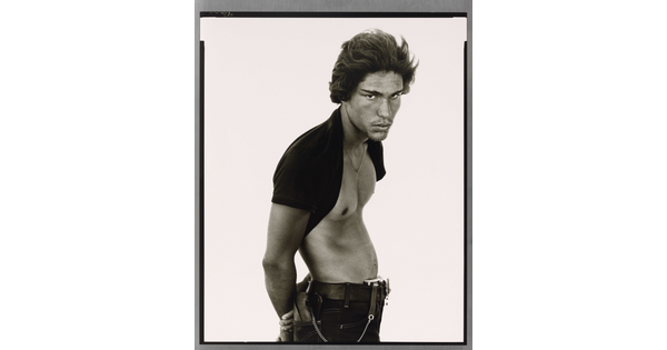 A black-and-white portrait photograph of a medium-skinned man standing sideways, posture in a subtle S-curve, arms behind his back, with a dark t-shirt pulled up behind his neck.