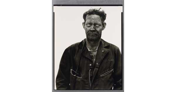 A black-and-white portrait photograph of a man in coveralls, hair sticking up, covered in dark coal dust.
