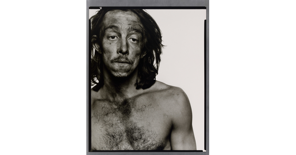 A black-and-white portrait photograph of a medium-skinned, shirtless man with long hair, and smudges of dark coal dust on his face.