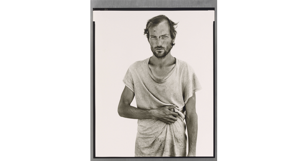 A black-and-white portrait photograph of a White man wearing a loose-fitting dirty white t-shirt, resting his arm across his stomach.