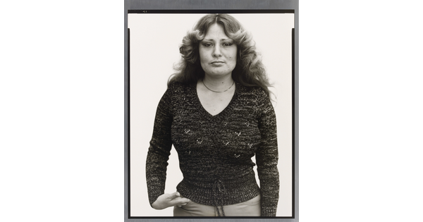 A black-and-white portrait photograph of a White woman with long hair curled back from her face, wearing a fitted dark sweater, and one hand in her pants pocket.