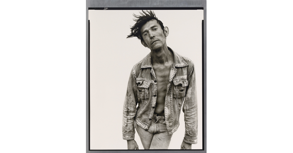 A black-and-white portrait photograph of a White man with dark hair that sticks up, wearing jeans and a jean jacket, and crossing his legs while bending forward.