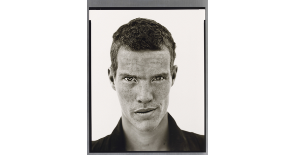 A black-and-white close-up photograph of a White man with short cropped hair, freckles across his face, wearing a dark collared shirt.