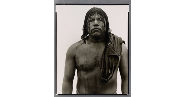 A black-and-white portrait photograph of a stocky, dark-skinned man with long, dark wet hair that drips down his shirtless torso.
