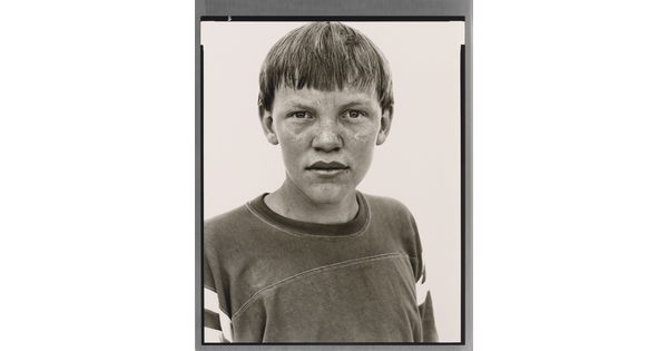 A black-and-white portrait photograph of a White teenage boy with a bowl haircut and freckles across his nose and cheeks, wearing an athletic t-shirt.