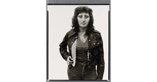 A black-and-white portrait photograph of a medium-skinned woman with dark, feathered hair wearing jeans, a jacket, V-neck top, and a heart necklace.