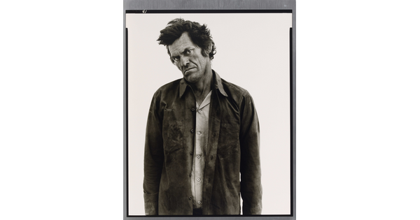 A black-and-white portrait photograph of a White man standing with his head cocked, frowning, and wearing two soiled shirts.