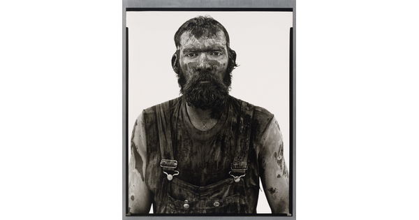 A black-and-white portrait photograph of a bearded man covered in mud and dirt, wearing dirty overalls, and looking straight at the camera.