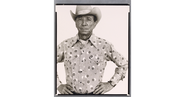 A black-and-white portrait photograph of an older White man wearing a cowboy hat, a button-down shirt, looking straight ahead with hands on hips.