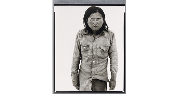 A black-and-white portrait photograph of a medium-skinned man with long hair, wearing dirty clothes, and squinting straight at the camera.