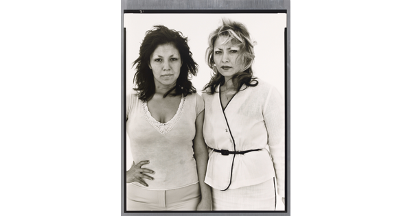 A black-and-white portrait photograph of two medium-skinned women, one in white pants and top with a hand on her hip and the other in a belted white suit, both looking intensely at the camera.