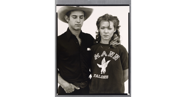 A black-and-white portrait photograph of a young White man wearing a cowboy hat, looking down, his hand affectionately around the neck of a young White woman.