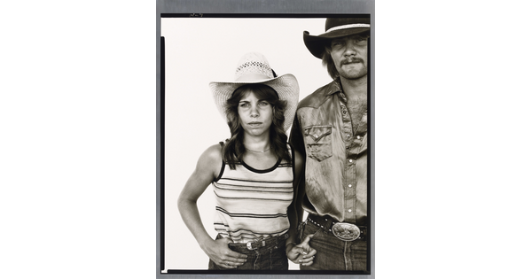 A black-and-white portrait photograph of a young White woman in a cowboy hat and striped tank top with one hand hooked into her jeans and the other hooked into the pocket of a White man in a cowboy hat and satin button-up shirt.