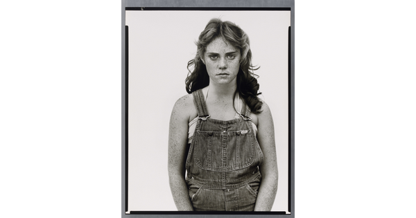A black-and-white portrait photograph of a young White girl with wavy hair, freckles on her face and arms, wearing denim overalls.