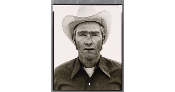 A black-and-white portrait photograph of a White man wearing a cowboy hat and collared shirt, with stubble on his face and chapped lips.
