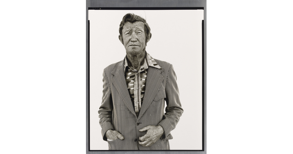 A black-and-white portrait photograph of an older White man with a worn face wearing a patterned, open-collar, button-down shirt under a chevron striped jacket.