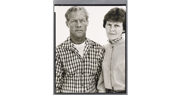 A black-and-white portrait photograph of a middle-aged White man wearing a checked button-down shirt and a middle-aged White woman in a striped high-collar blouse standing shoulder to shoulder.