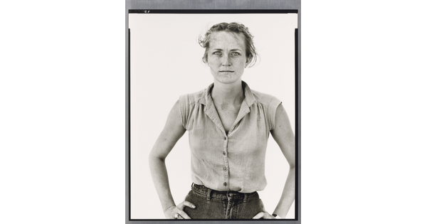 A black-and-white portrait photograph of a young White woman in a sleeveless blouse, hair pulled back from her face, and hands on her hips.