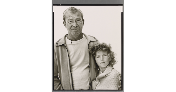 A black-and-white portrait photograph of an older White man in a t-shirt and cardigan with his arm around a young White girl with curly hair and a blanket wrapped around her shoulders.
