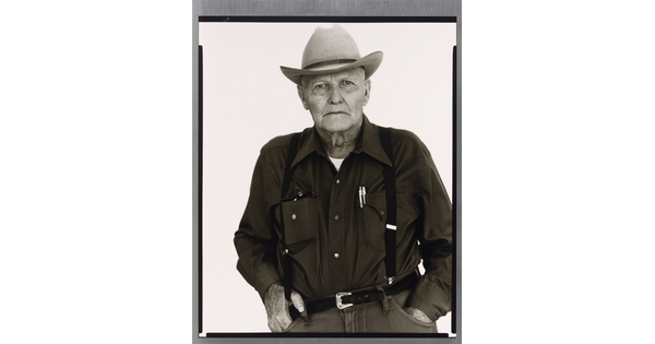 A black-and-white portrait photograph of an older White man wearing a cowboy hat, button down shirt, pants with suspenders and his hands in his pockets