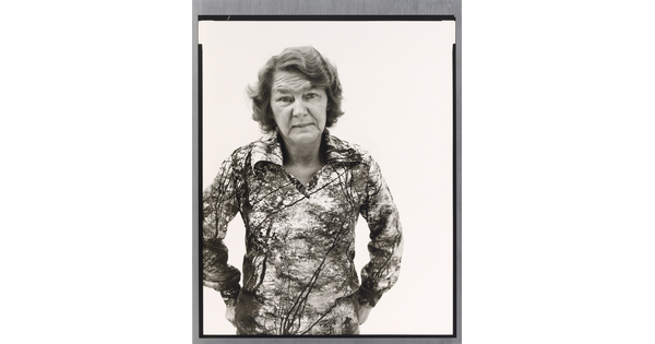 A black-and-white portrait photograph of an older White woman with wavy hair wearing a patterned polo shirt, hands hooked into her back pants pockets.