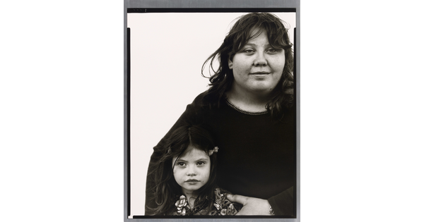 A black-and-white portrait photograph of a medium-skinned woman with dark hair holding close to her a young medium-skinned girl with long dark hair in barrettes.