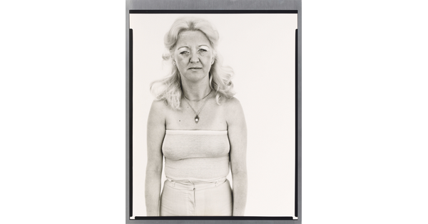 A black-and-white portrait photograph of a White woman with long, bleach-blonde hair in a white tube top and pants.