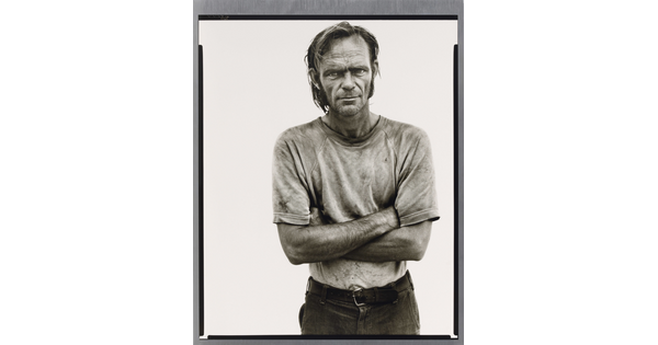 A black-and-white portrait photograph of a White man with unkempt hair in a dirty tee-shirt standing with arms crossed.